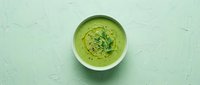 Fresh green cream soup in a white bowl, topped with microgreens and black pepper, on a light mint textured background. Top view.