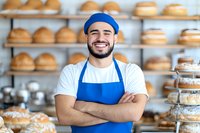 Cheerful young Hispanic male baker in blue uniform standing with crossed arms at bakery counter surrounded by fresh bread and pastries on display shelves.