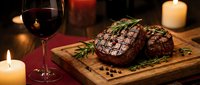 Grilled beef steak with herbs on wooden board, red wine glass and candles creating romantic dinner atmosphere for fine dining restaurant menu.