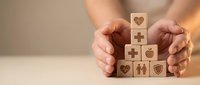 Hands protecting wooden blocks with healthcare and insurance icons including heart, medical cross, family and shield symbols on neutral background.