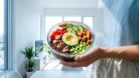 Young adult male with medium skin tone holding a balanced bowl meal featuring quinoa, fresh vegetables, and protein, displayed in bright natural light from window, suitable for health, nutrition, wellness, and lifestyle content.