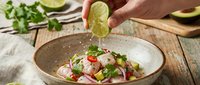 Hand squeezing fresh lime juice over vibrant ceviche salad with seafood, vegetables and herbs in ceramic bowl for cooking and food preparation.