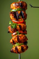 Grilled chicken kebab with bell peppers and tomatoes on metal skewer, garnished with fresh herbs against green background. Juicy meat pieces show charred grill marks.