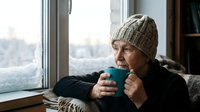 Senior Caucasian woman in knit beanie enjoying warm beverage by window on cold winter day, cozy indoor comfort and seasonal relaxation at home.