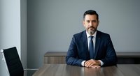 Mature male professional with dark hair and facial hair wearing navy business suit and tie, seated at wooden desk in modern office environment with gray walls and contemporary furniture, suitable for corporate, business leadership, and professional services marketing.