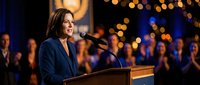 Professional businesswoman speaking at podium during corporate event with warm bokeh lights and audience in background.