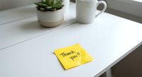 Yellow sticky note with thank you message on white wooden desk beside coffee mug and succulent plant for office gratitude and workplace appreciation concepts.