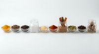 Assorted spices, herbs and ingredients arranged in glass bowls and jars on white background for cooking and culinary preparation.