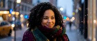 Young African American woman with curly hair wearing colorful scarf smiling on snowy city street at night with warm lights and bokeh background.