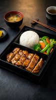 Traditional Japanese bento box with teriyaki chicken, steamed rice and vegetables served with miso soup and tea on dark slate surface for authentic dining.