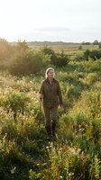 Middle-aged Caucasian woman in olive green outdoor clothing walking through wildflower meadow at golden hour with rolling countryside landscape background.