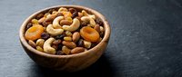 Wooden bowl filled with mixed nuts and dried fruits including cashews, almonds, raisins on dark slate surface for healthy snacking and cooking.