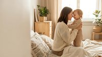 Young Caucasian mother in beige sweater holding smiling toddler daughter on bed in bright modern bedroom with plants and natural morning light streaming through window.