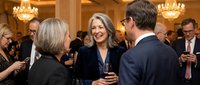 Mature woman with gray hair and glasses in dark blazer smiling while conversing with man in navy suit at formal event in elegant ballroom with chandeliers and guests networking.