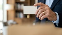 Mature male professional in dark business suit holding blank white card in office environment with blurred background for corporate messaging and branding purposes.