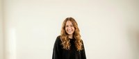 Young Caucasian woman with long curly blonde hair smiling warmly in black sweater against neutral background for lifestyle and portrait photography.