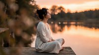 Young Asian woman in white clothing meditating peacefully on wooden dock at sunset by calm lake water with warm golden light reflecting on surface.