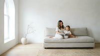 Young mother and toddler daughter reading book together on modern beige sofa in minimalist living room with natural light from large window.