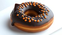 Fresh donut covered with dark chocolate glaze and decorated with orange candy and black sprinkles on white plate, close up view of sweet dessert food.