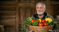 Senior man with white hair and beard, Caucasian ethnicity, farmer or gardener profession, holding wicker basket filled with fresh vegetables including tomatoes, peppers, and leafy greens in rustic wooden setting.