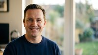 Middle-aged Caucasian man smiling warmly in casual navy sweater at home with natural window light creating friendly approachable portrait for lifestyle content.