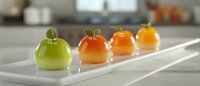 Colorful fruit-shaped desserts arranged in a row on white rectangular plate, showcasing gradient from green apple to orange varieties in modern kitchen setting.