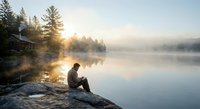 Young man sitting on rocky shore drinking coffee at misty lake during golden sunrise with cabin and pine trees reflecting in calm water.