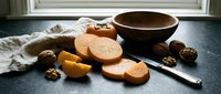 Sliced ripe persimmons with walnuts, a wooden bowl, and a knife on a dark stone surface with a linen napkin in natural light.