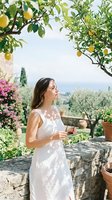 Young woman with long dark hair and Mediterranean ethnicity standing in a lemon orchard garden, wearing a white sleeveless dress, holding a wine glass, surrounded by blooming pink flowers and lush green foliage with scenic countryside landscape in background.