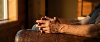 Elderly man's weathered hands clasped together in contemplative prayer or meditation, sitting peacefully in warm golden sunlight streaming through window.