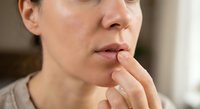Middle-aged Caucasian woman touching lips with finger in contemplative gesture, showing concern or worry in soft natural lighting for healthcare concepts.