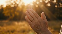 Elderly hands raised in prayer or gratitude during golden hour sunset, spiritual moment of reflection and faith in warm natural light outdoors.