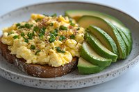 Creamy scrambled eggs on toasted sourdough bread with fresh sliced avocado, garnished with chives and black pepper, served on ceramic plate. Perfect breakfast.