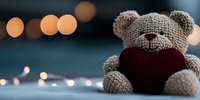 Soft plush teddy bear holding red heart cushion against blurred bokeh lights background, romantic Valentine's day composition on blue textile surface.