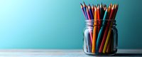 Colorful wooden pencils standing in clear glass jar against turquoise blue wall on wooden table surface, minimalist education and creativity concept.