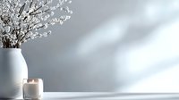 White ceramic vase with delicate cherry blossom branches and scented candle in glass holder on white surface against light gray wall with soft natural shadows.