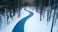 Winding blue road curves through snowy winter forest landscape with tall trees, aerial drone view capturing pristine wilderness and natural path perspective.