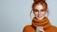 Young redhead woman in glasses and cozy orange knit scarf smiling while holding smartphone against light gray background. Casual winter lifestyle portrait.