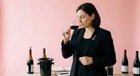 Professional female sommelier in black suit tasting red wine at elegant tasting event with multiple bottles on pink background for hospitality industry.