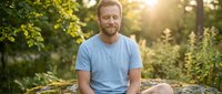 Middle-aged Caucasian man with reddish-blonde beard wearing light blue t-shirt sitting outdoors on rock surrounded by green forest trees during golden hour sunset, relaxed casual portrait.