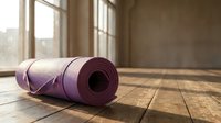 Purple yoga mat rolled up on wooden floor in bright studio room with natural sunlight streaming through windows for fitness and wellness practice.