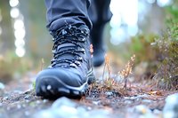Close-up of black hiking boot on forest trail with dried plants and bokeh background, showcasing outdoor adventure gear and nature exploration details.
