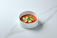 Fresh gazpacho soup garnished with cucumber slices and herbs in white ceramic bowl on marble surface, cold summer appetizer served restaurant style.