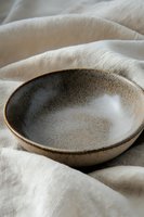 Handcrafted ceramic bowl with rustic speckled glaze on natural linen fabric background, showcasing artisanal pottery details in soft natural lighting.