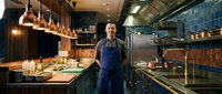 Confident male chef in blue apron standing in a modern professional restaurant kitchen with copper accents and pendant lights.