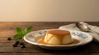 Creamy caramel flan dessert with golden syrup on decorative plate with mint garnish and coffee beans on rustic wooden table for culinary photography.
