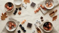 Autumn flat lay with artisan chocolates, coffee cups, fall leaves and cozy seasonal treats arranged on neutral background for food styling and design.
