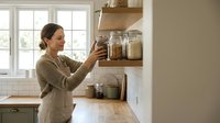 Young woman organizing glass jars on wooden kitchen shelves in modern home interior for meal prep and pantry storage solutions.