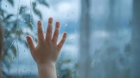 Child hand pressed against rainy window glass with water droplets, expressing longing and melancholy on cloudy day indoors.