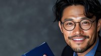 Young Asian businessman with glasses and modern hairstyle showing bright confident smile while holding blue folder against dark background.